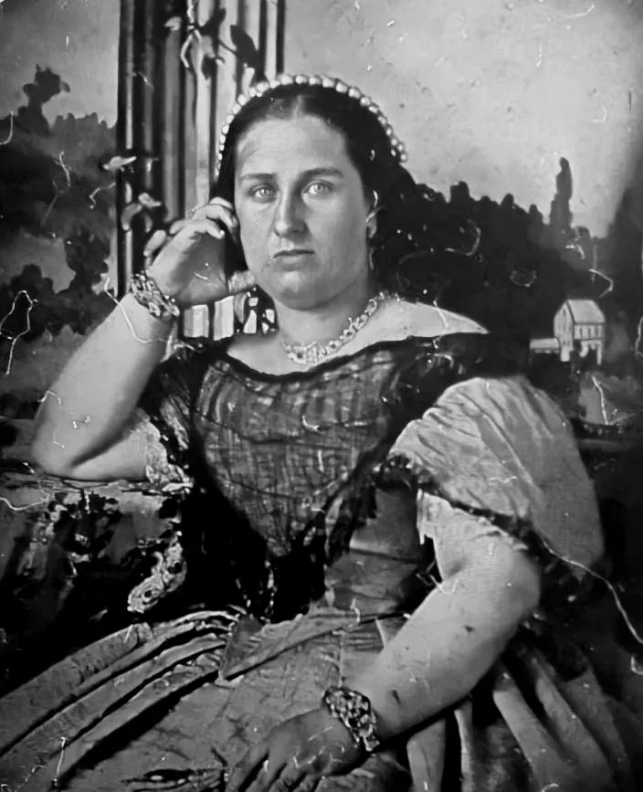 A faded early photograph of a heavy-set woman with dark hair parted in the centre and a pearl diadem, resting her head on one hand. She wears a dark dress with a wide neckline, a necklace and broad bracelets, in front of a painted backdrop.