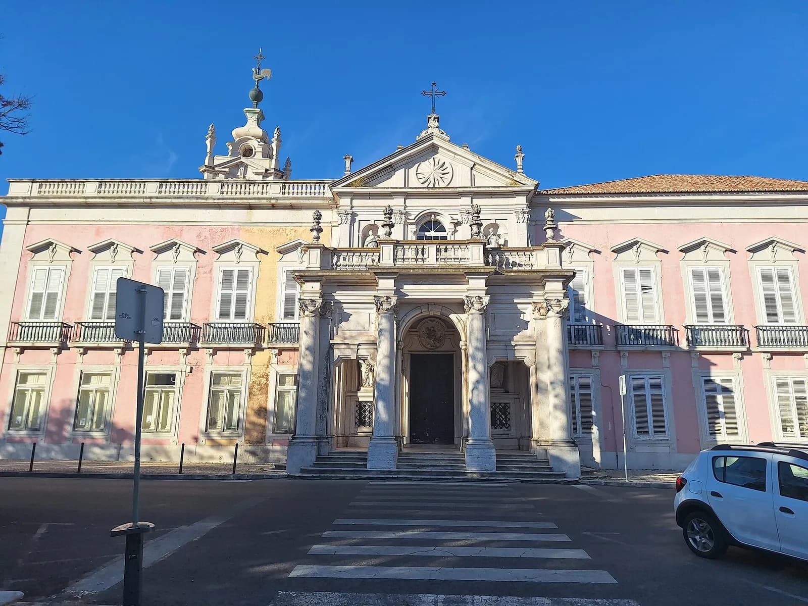 A long pink palace façade with white stone window frames and balconies, a marble porch with columns and a pediment topped by a cross in the centre, and a small domed bell tower on the left, under a clear blue sky.