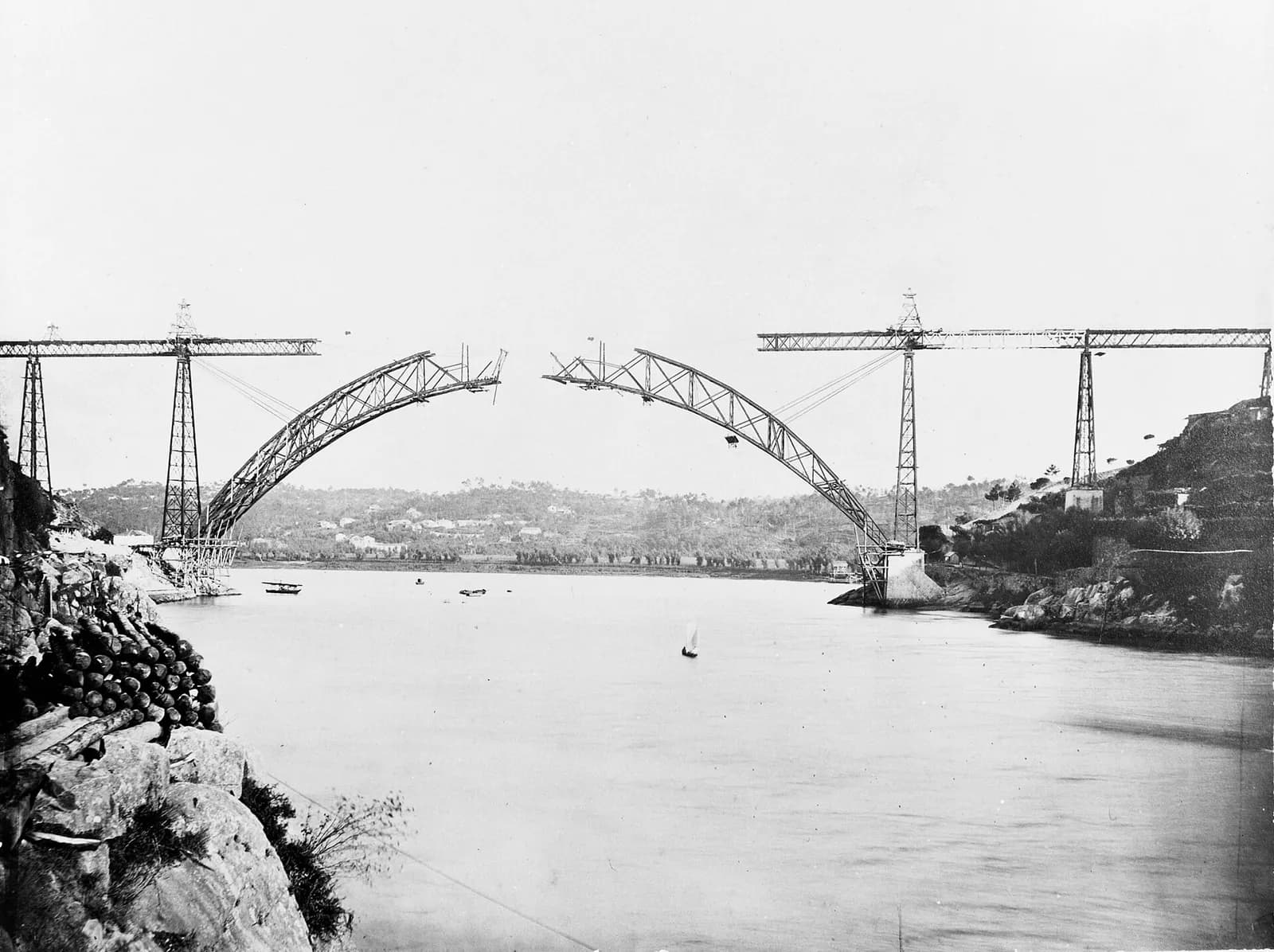 Early photograph of a wide river between rocky banks. From each side a curved lattice half-arch reaches out over the water towards the other, almost meeting in the middle. Lines of cable run from the tips of the half-arches back to tall iron piers carrying a straight deck on each bank.