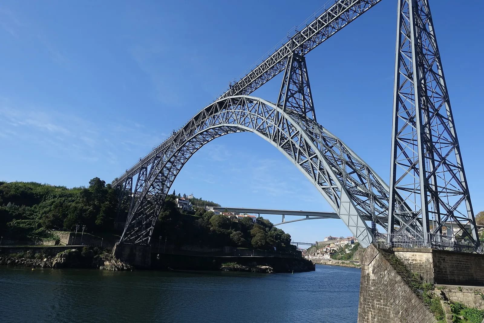 A tall grey wrought-iron arch, made of two latticed ribs, springs from a stone abutment on the right across a blue river. A straight lattice deck runs over the top on slender iron piers. Wooded banks rise on either side, and a flat concrete road bridge crosses the river further downstream.