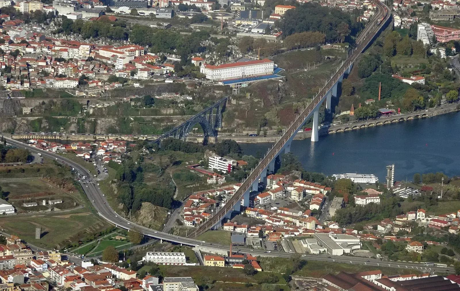 Aerial photograph of a river valley lined with houses. On the left a slender grey iron arch bridge crosses the river; to its right, a long straight concrete railway bridge on tall white piers runs diagonally across the valley and up into the city.
