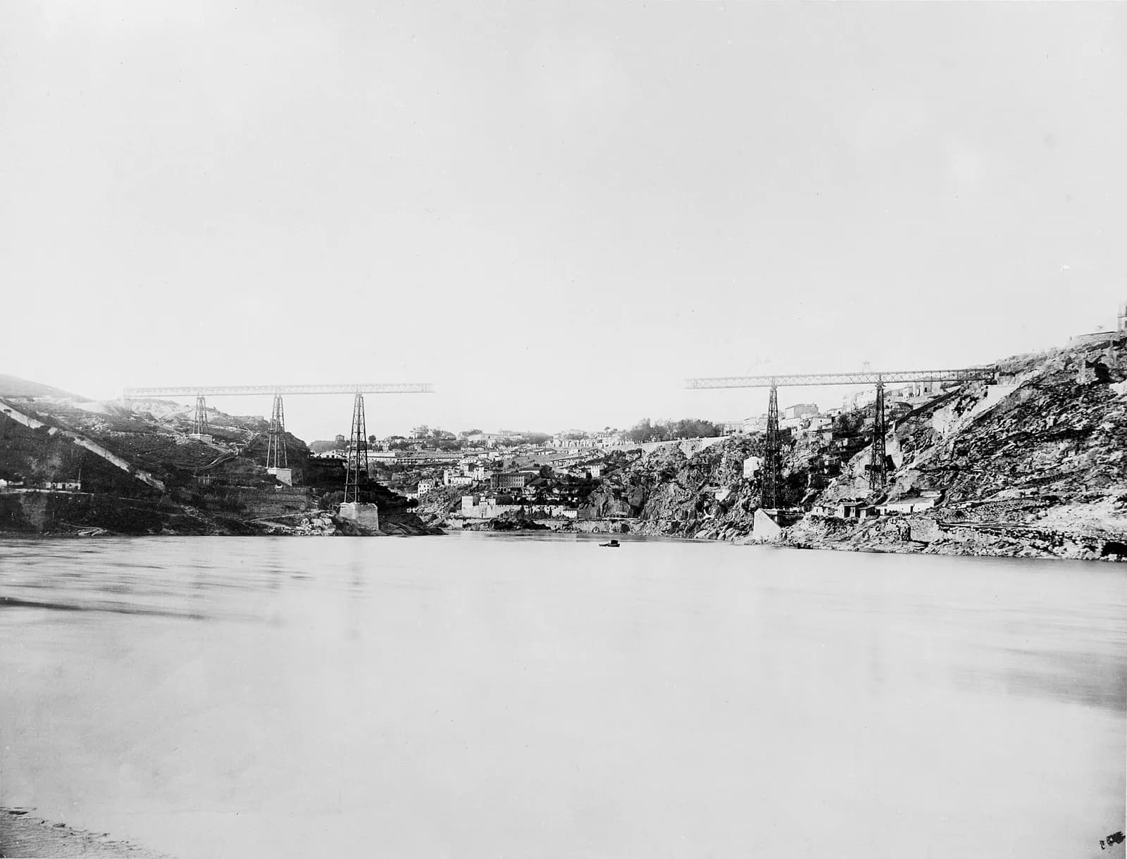 Early photograph taken from the water's edge of a wide, calm river between steep rocky banks. On each bank, tall lattice iron piers on masonry bases carry a straight lattice girder that stops in mid-air over the river: three piers on the left bank, two on the right. Houses climb the hill in the distance.
