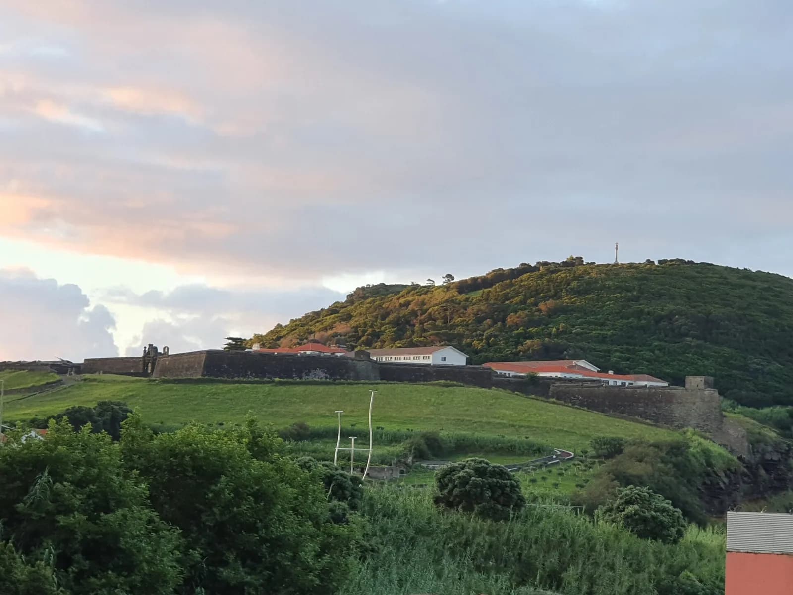 Long dark stone ramparts with low white buildings and red roofs stretch along the foot of a green, wooded hill under a pink-tinged sky. Fields and shrubs fill the foreground.