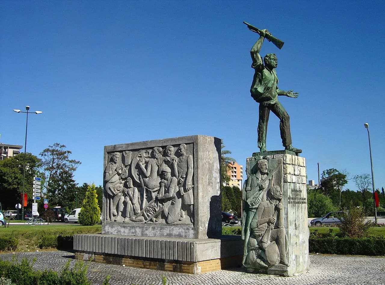A stone monument on a paved island under a blue sky. On the left a block carved in relief shows a crowd of working men and women; on the right a squared stone pillar, carved with two workers in relief, carries a dark green statue of a man striding forward and holding a rifle high above his head.