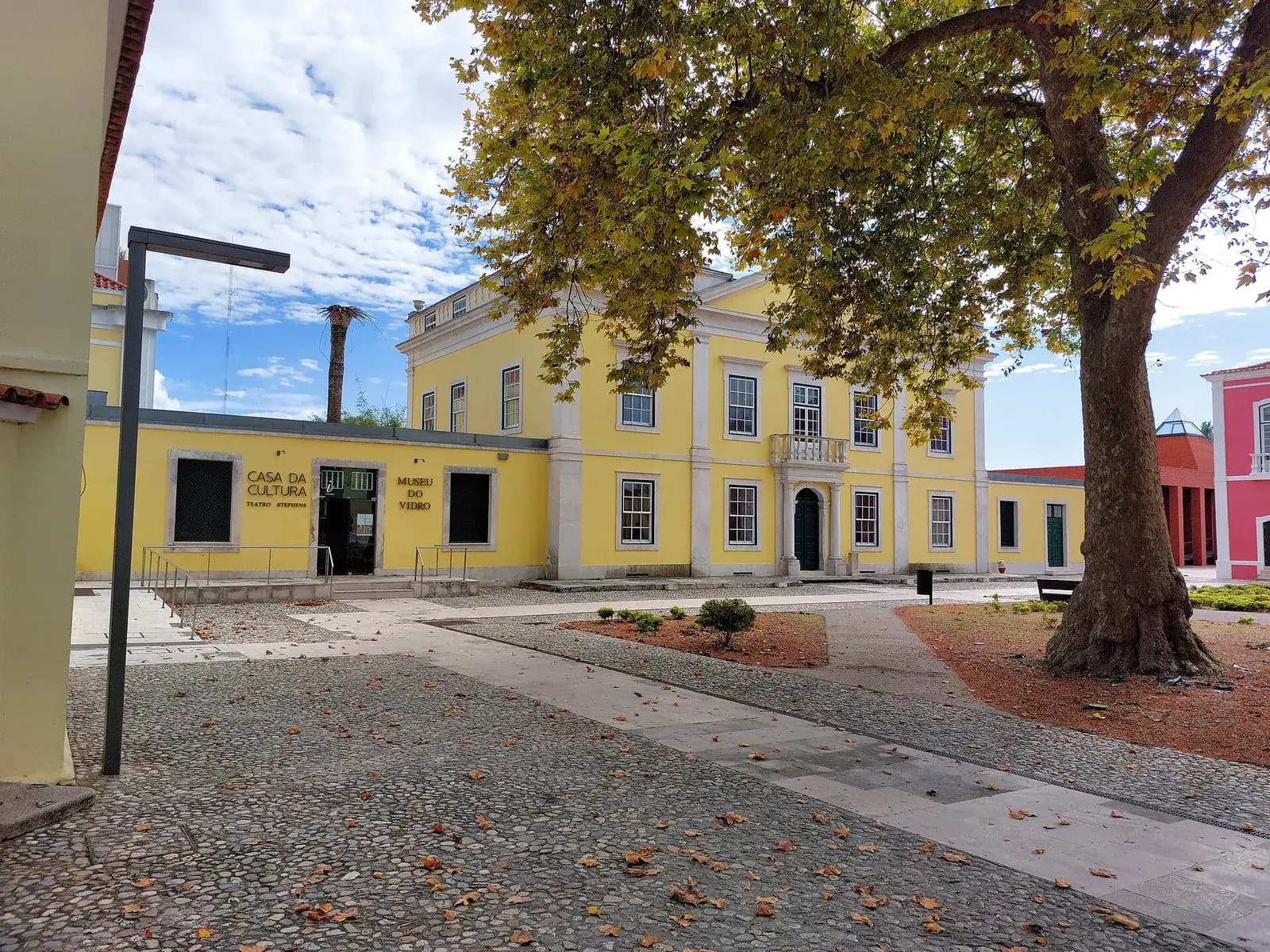 A yellow two-storey neoclassical mansion with white stone pilasters, a small balcony over the central door and a low yellow wing to the left lettered Casa da Cultura and Museu do Vidro. A large plane tree shades a cobbled square in front.