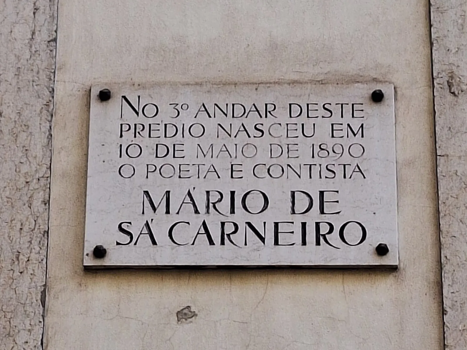 A white marble plaque with black capital letters fixed by four bolts to a pale building wall, reading in Portuguese that the poet and short-story writer Mário de Sá Carneiro was born on the third floor of this building on 10 May 1890.