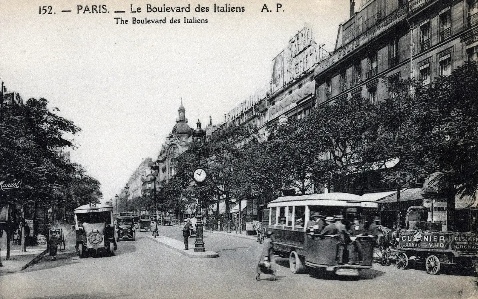 An old black-and-white postcard of a wide Paris boulevard lined with trees and tall stone buildings. Motor buses, a motor cab and a horse-drawn cart share the road, a street clock stands on a traffic island, and pedestrians cross in the foreground.