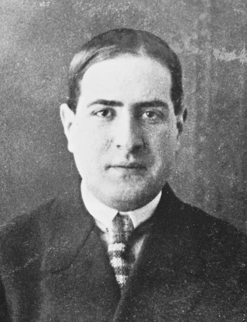 Black-and-white head-and-shoulders portrait of a heavy-set young man with dark hair combed back and parted, a white collar, a checked tie and a dark jacket, looking at the camera.