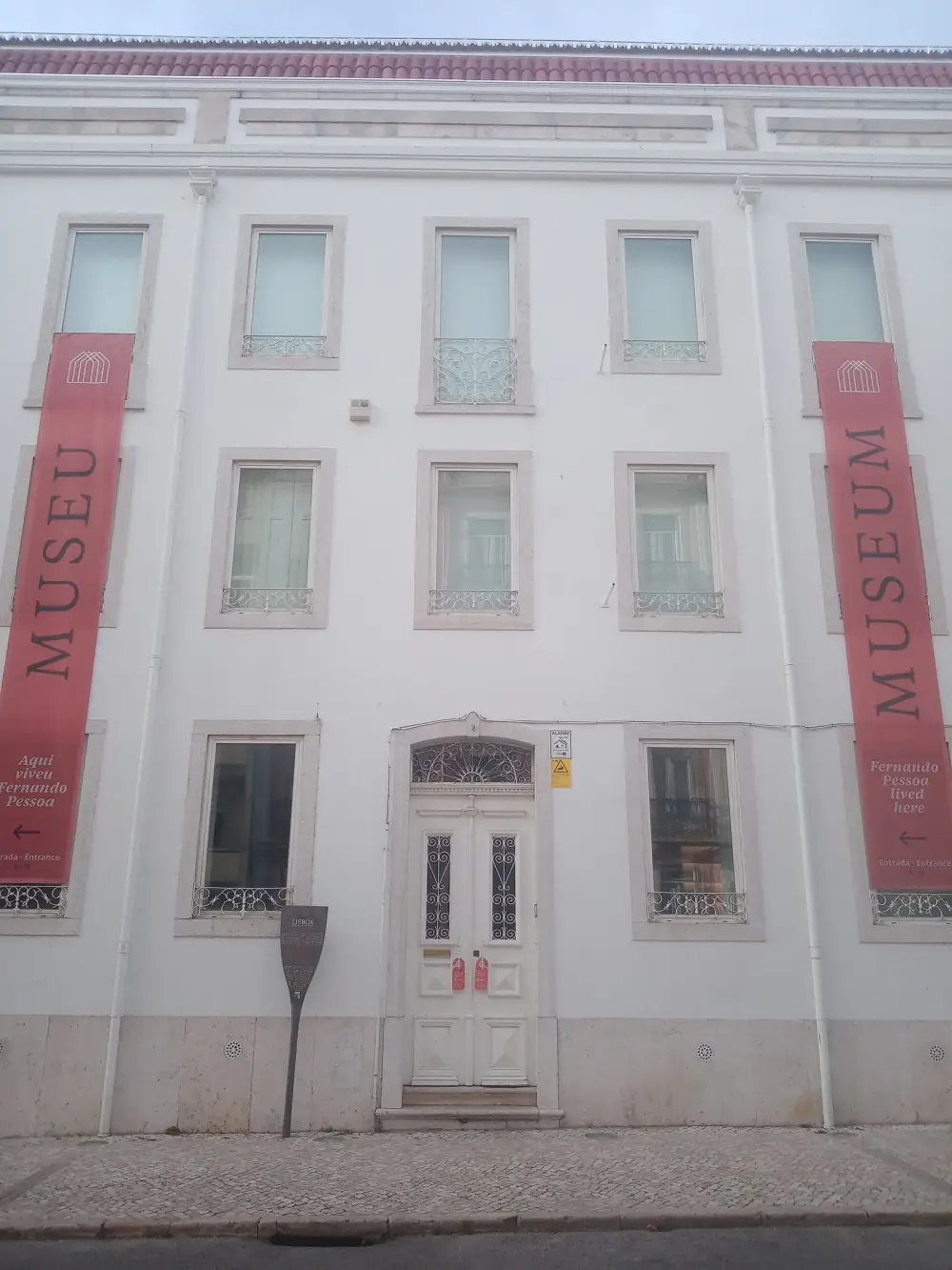 A plain white three-storey building with a panelled double door in the centre. Long red banners hang on either side, one reading Museu, Aqui viveu Fernando Pessoa and the other Museum, Fernando Pessoa lived here.