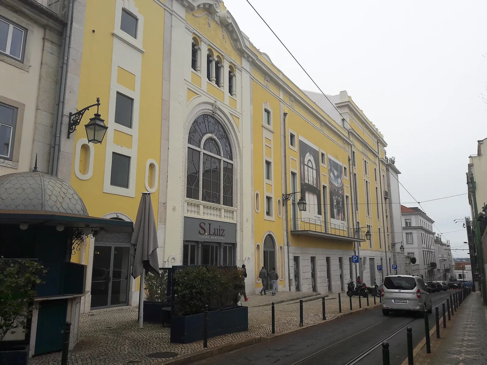 The yellow and white façade of a theatre on a sloping Lisbon street, with a tall arched window above an entrance sign reading S. Luiz Teatro Municipal, and tram rails in the street.