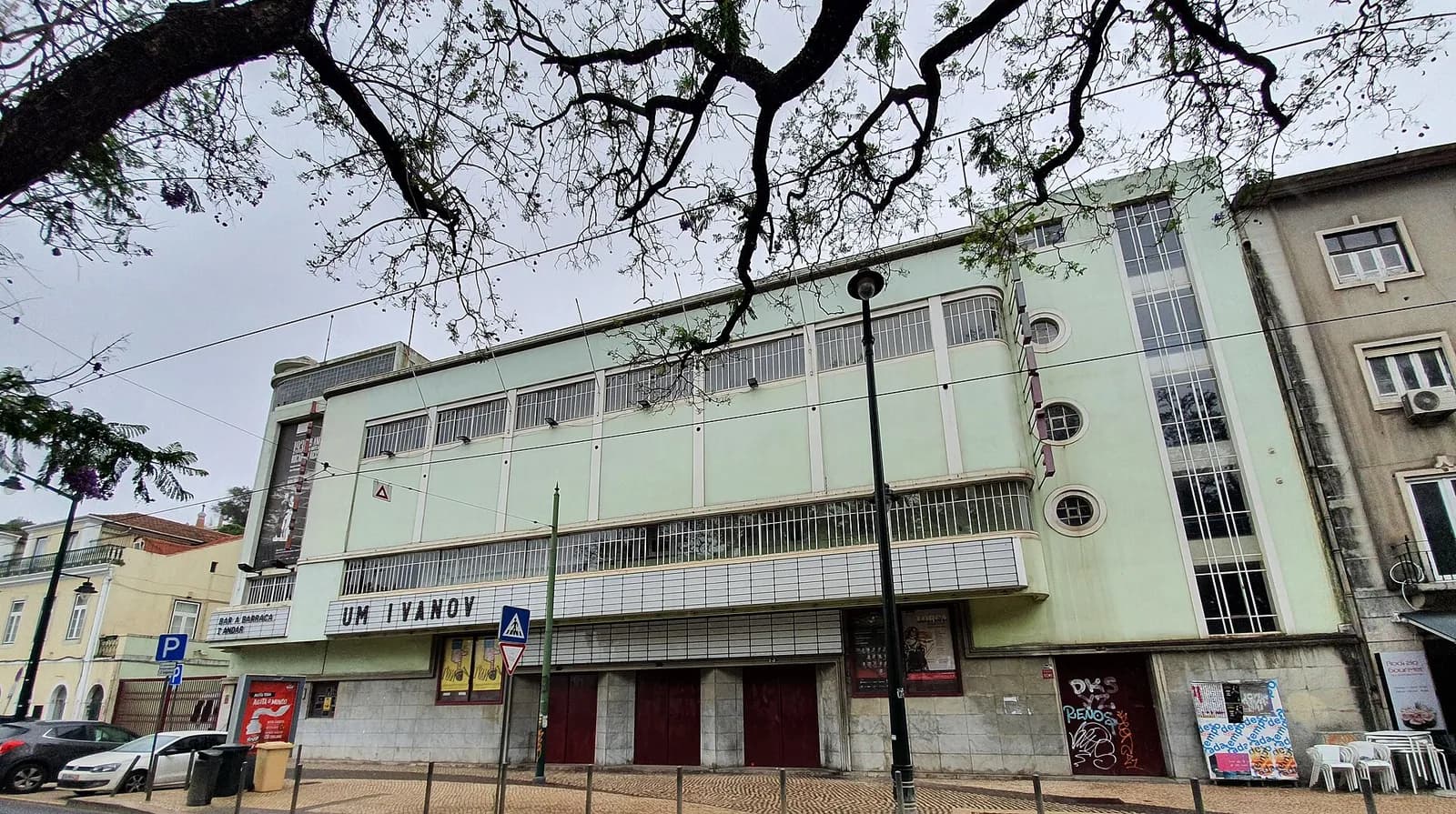 A pale green modernist cinema building on a square, seen through the branches of a tree, with a long sign over the entrance reading Um Ivanov and a smaller one at the corner reading Bar A Barraca.
