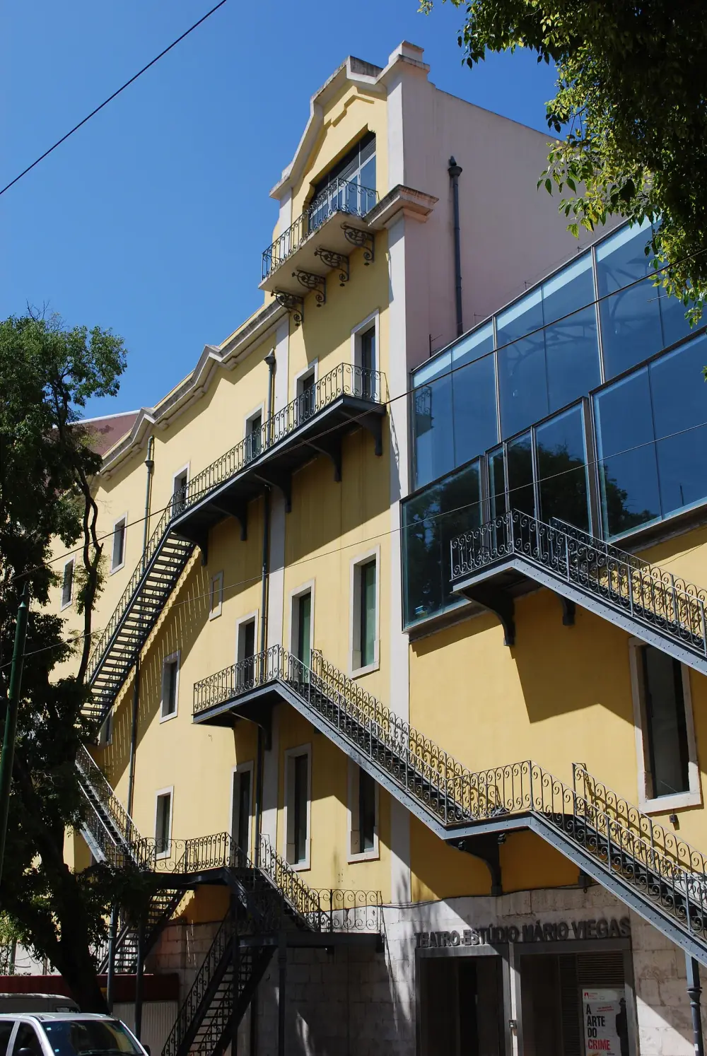 The tall yellow side wall of a theatre with grey wrought-iron fire escapes zigzagging down it and a glass extension at the top right. Over a ground-floor doorway a sign reads Teatro-Estúdio Mário Viegas.