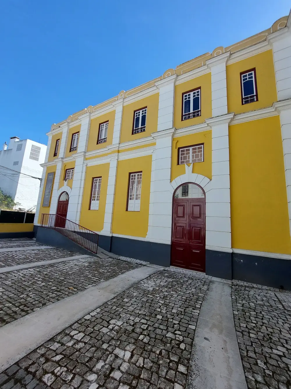 A two-storey yellow and white theatre building with dark red arched doors, seen at an angle across a cobbled square under a blue sky.