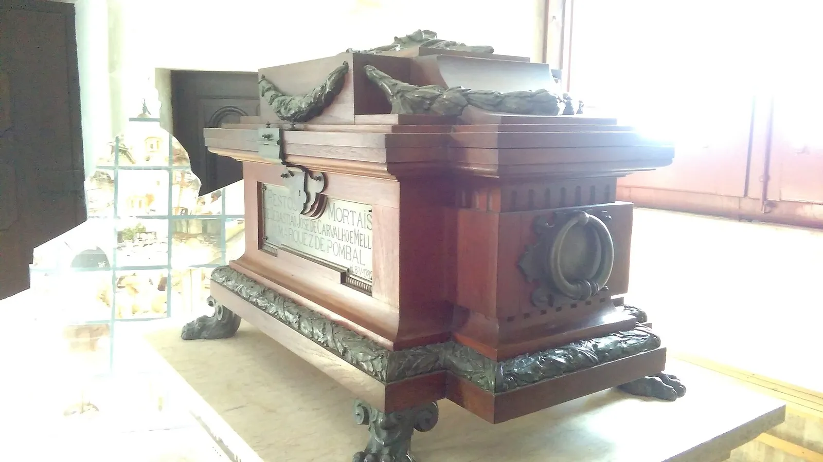 A polished dark wooden urn with bronze laurel garlands on the lid, bronze lion's-paw feet and a large bronze ring on its end, standing on a stone plinth in a bright church. A silver plaque on the side reads Restos mortais de Sebastião José de Carvalho e Mello, Marquez de Pombal, with the date 8-V-1782 in a corner.