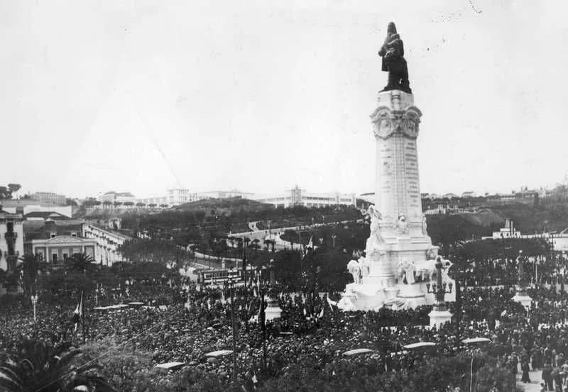 Black-and-white photograph of a huge crowd filling a square around a tall white column topped by a bronze statue of a man with a lion beside him, with gardens and buildings on the hill behind.
