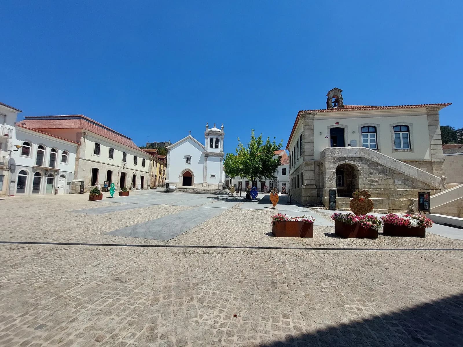 A wide cobbled square under a clear blue sky. On the left is a long two-storey building with arched ground-floor doors; in the centre a small white church with a bell tower; on the right a stone building with an outside staircase and a bell gable. A castle stands on the hill behind the left-hand buildings.