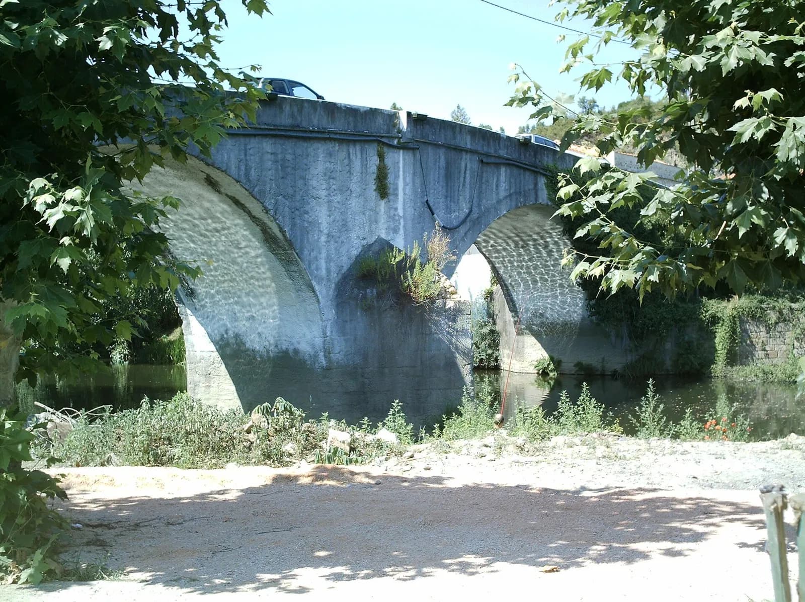 A stone bridge with two wide round arches over a calm river, framed by plane-tree leaves, with cars on the roadway above and a gravel bank in the foreground.