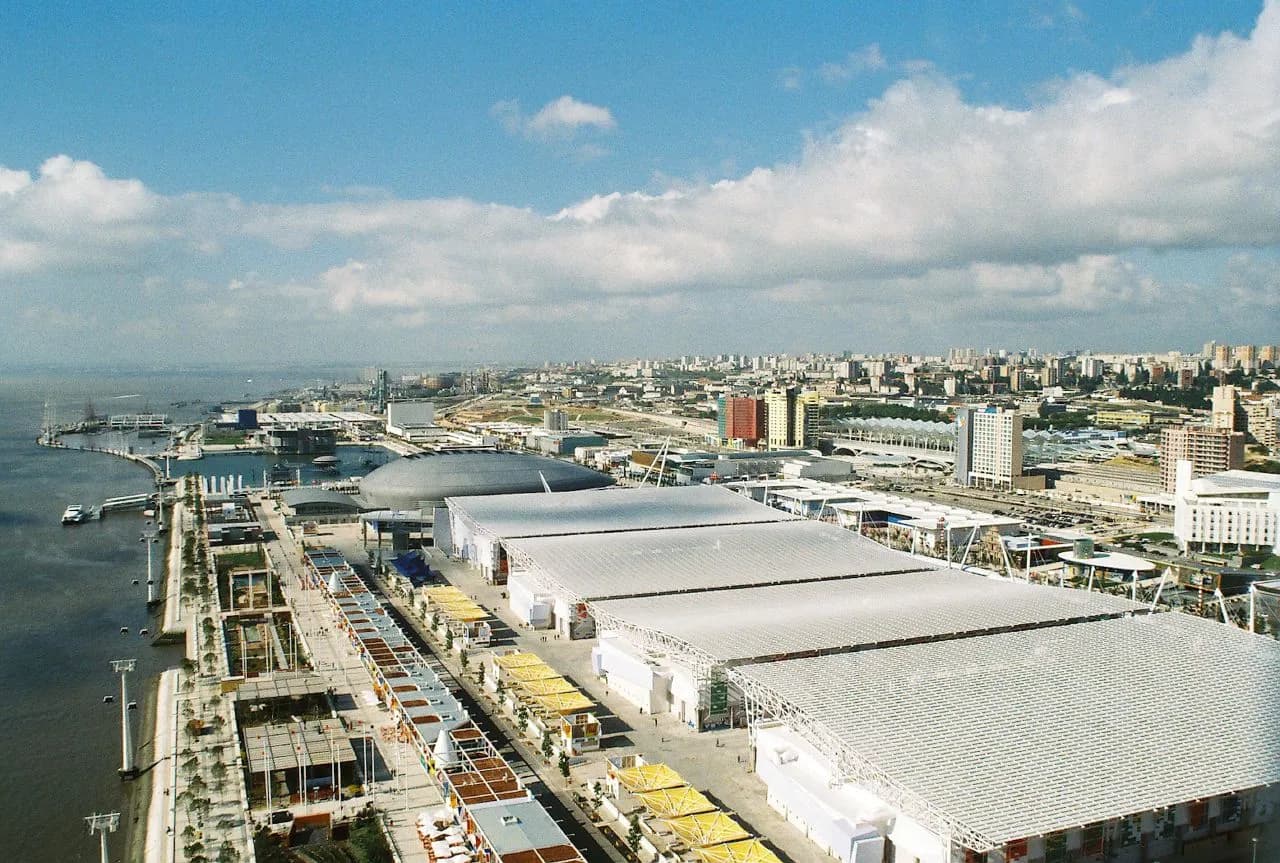 Aerial view of a riverside exhibition ground under a blue sky with white clouds. Long, low halls with pale translucent roofs fill the foreground beside a promenade lined with stalls and yellow awnings. Beyond them a grey oval dome sits near the water, and the apartment blocks of eastern Lisbon spread to the horizon on the right.