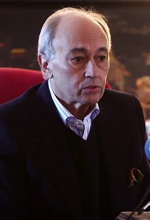 A grey-haired man in a dark jacket, dark jumper and striped shirt with a handkerchief in his breast pocket speaks into a microphone, seated in front of a red chair.