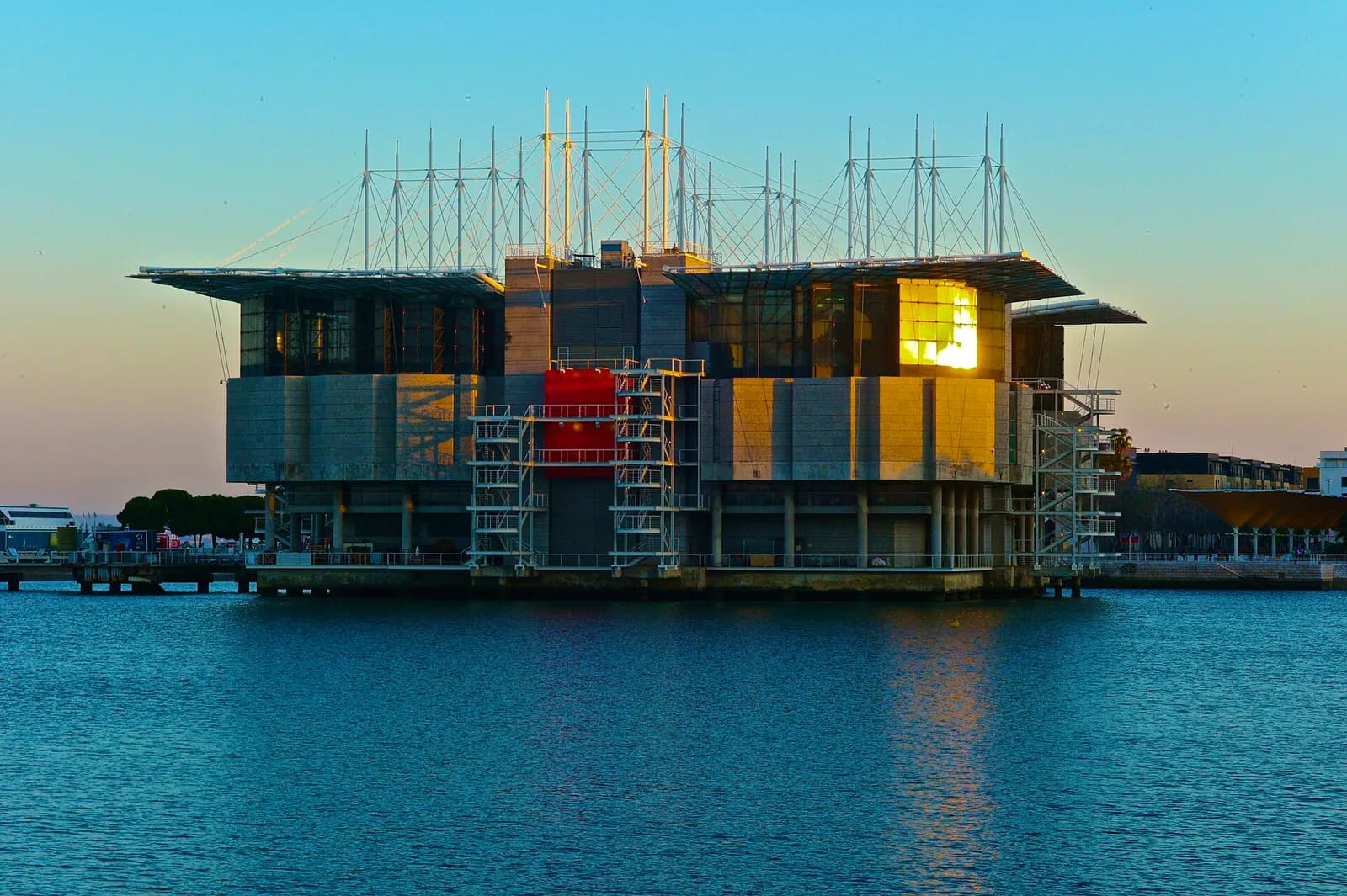 A boxy grey building with glass upper storeys and a red panel stands on a platform in calm water at dusk. Masts and cables rise from its roof, and the low sun lights one window gold.