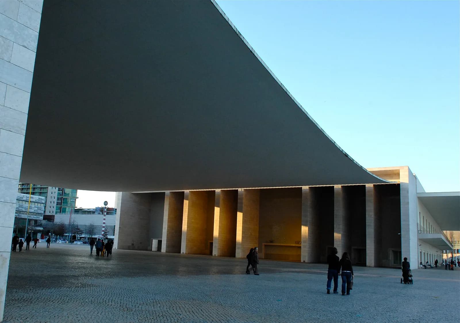 A vast, thin concrete canopy curves down over a paved square, held between a stone-clad wall on the left and a colonnade of tall stone piers lit by low sun. A few people walk across the square beneath it.