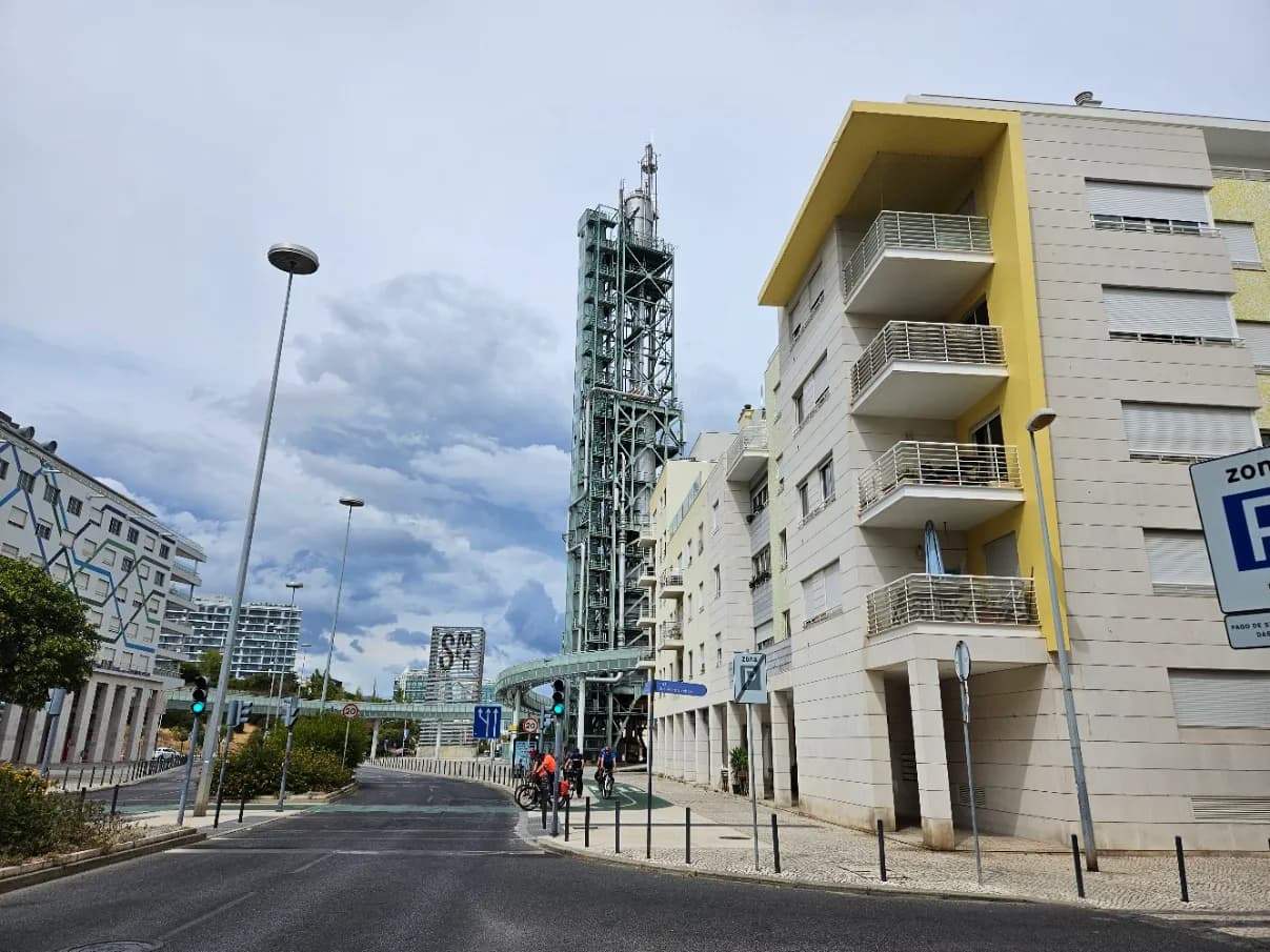 A tall green steel lattice tower wrapped in pipes and ladders rises between a curving footbridge and a pale apartment block with yellow trim, on a residential street under a cloudy sky.