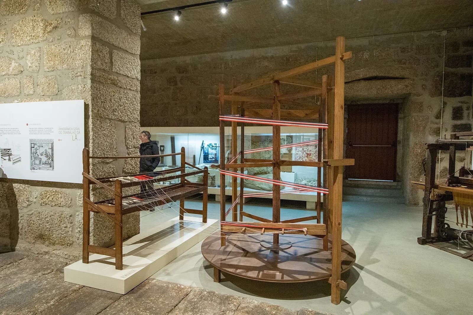 A museum room with rough granite walls and a stone floor, displaying wooden textile equipment: a tall rotating frame strung with red threads, a low wooden frame and, at the right, part of a loom.