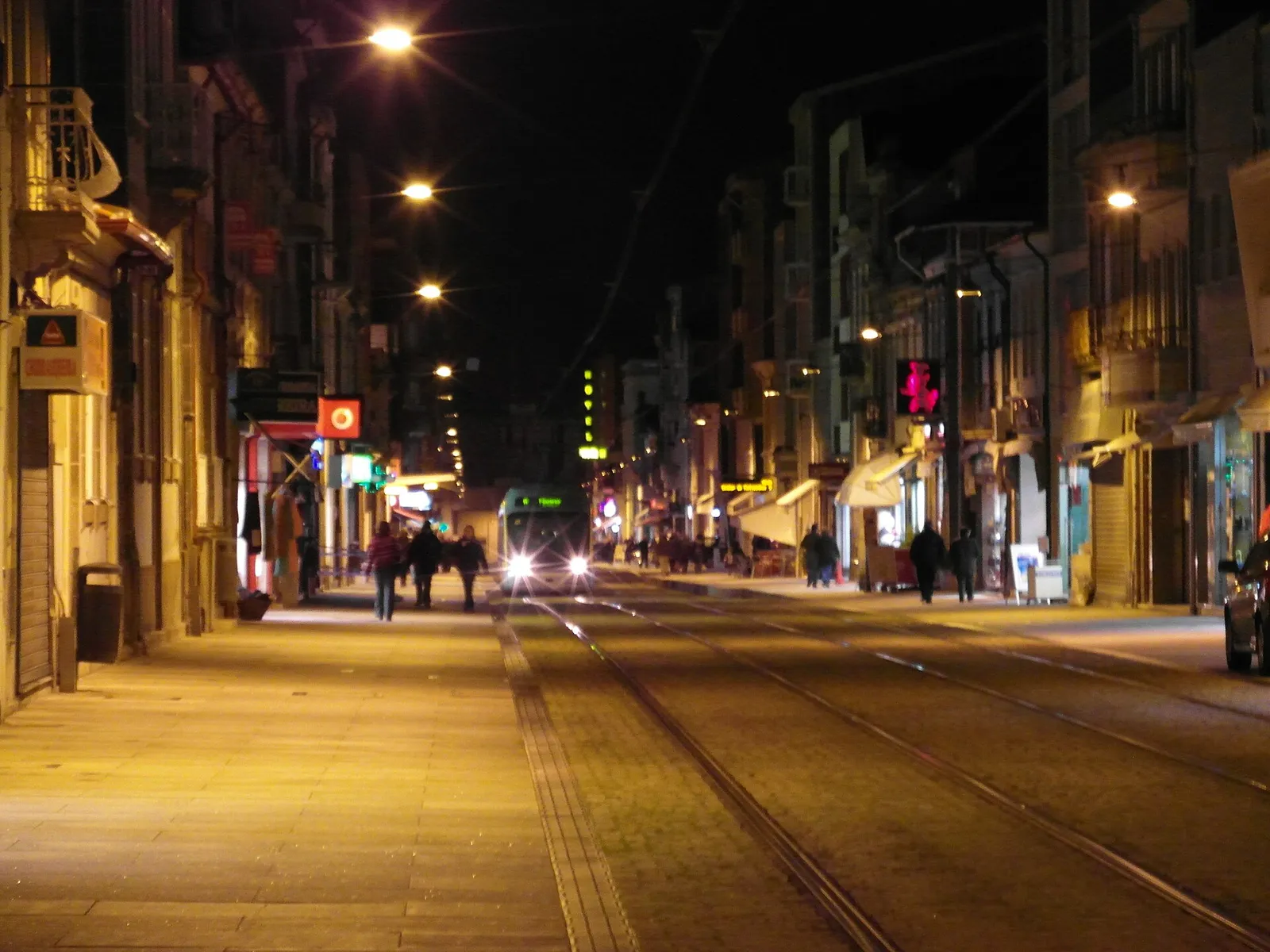 A narrow street at night lined with lit shopfronts and old houses, with two pairs of rails set into the cobbled roadway. A light-rail train with its headlights on approaches in the distance, and pedestrians walk on the pavements.