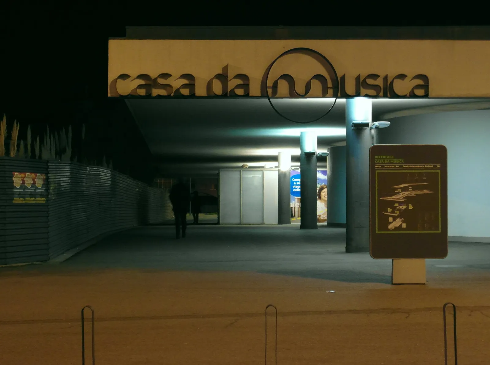 A low concrete canopy at night with large lettering reading casa da música, a big circle drawn over the letters. Beneath it are lit columns and a sign with a map of the interchange, and a dark figure walks into the covered space.