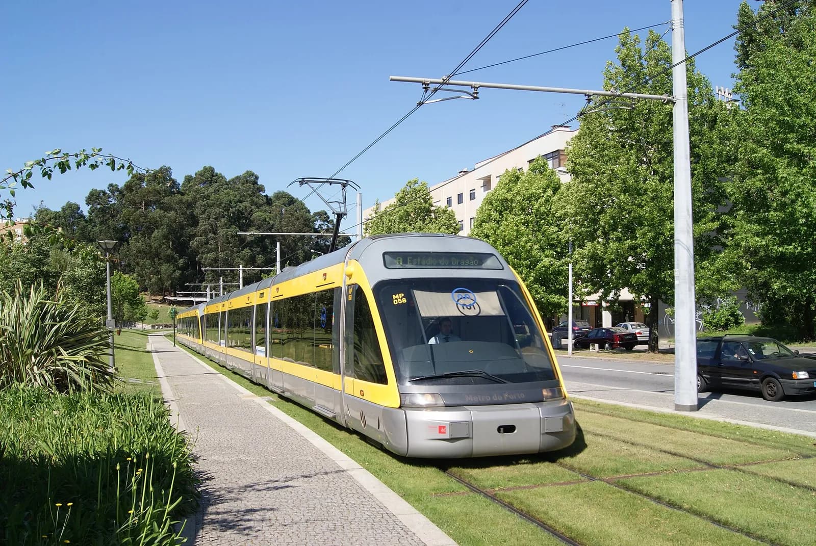 A grey and yellow light-rail train with the destination A Estádio do Dragão runs on grass-covered tracks beside a paved footpath and a road, under overhead wires, with trees and a block of flats behind it on a sunny day.