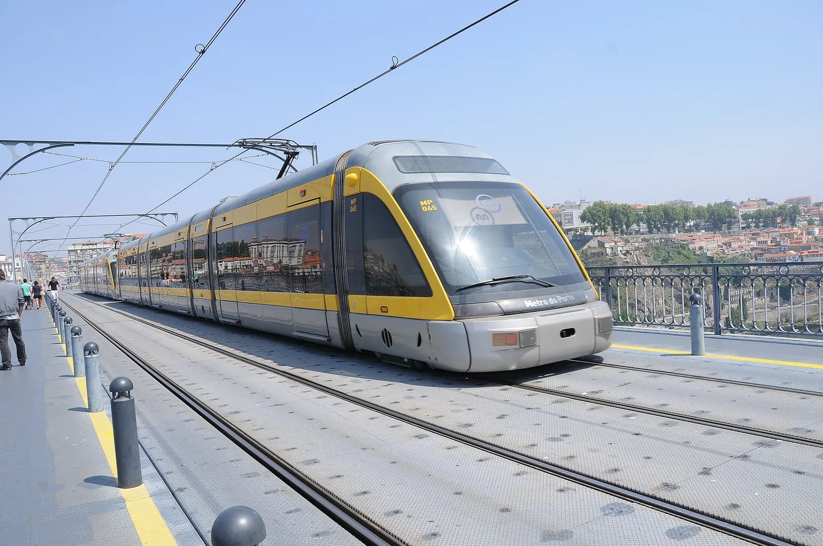 A grey and yellow Metro do Porto train on a metal bridge deck with two tracks under overhead wires, a railed footway with bollards to the left and houses on a hillside across the river in the background.