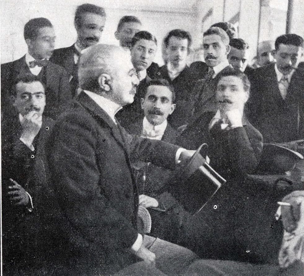 Black-and-white photograph of a white-haired man in a dark coat, seen in profile and holding a top hat, seated among a crowd of younger men with moustaches who look on.