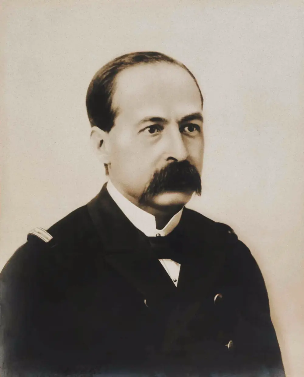 Sepia studio portrait of a balding man with a thick dark moustache, wearing a dark naval uniform jacket with a high white collar and bow tie.