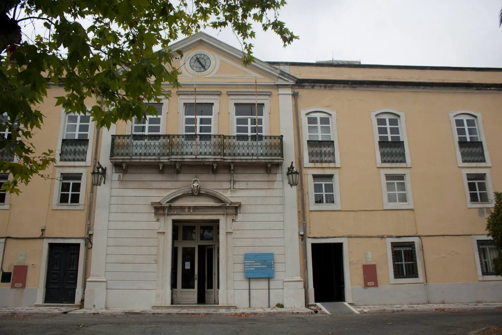Three-storey ochre building with white stone trim, a central bay with a clock in its pediment and a wrought-iron balcony above the main door, partly framed by tree branches.