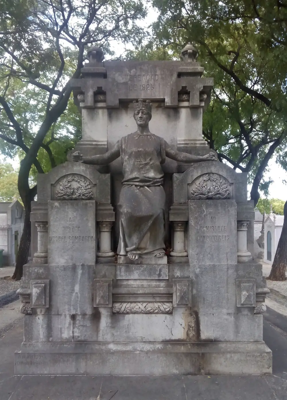 Weathered stone monument with a seated female figure in a turreted crown holding out both arms, flanked by two inscribed slabs under carved laurel wreaths, with trees behind.