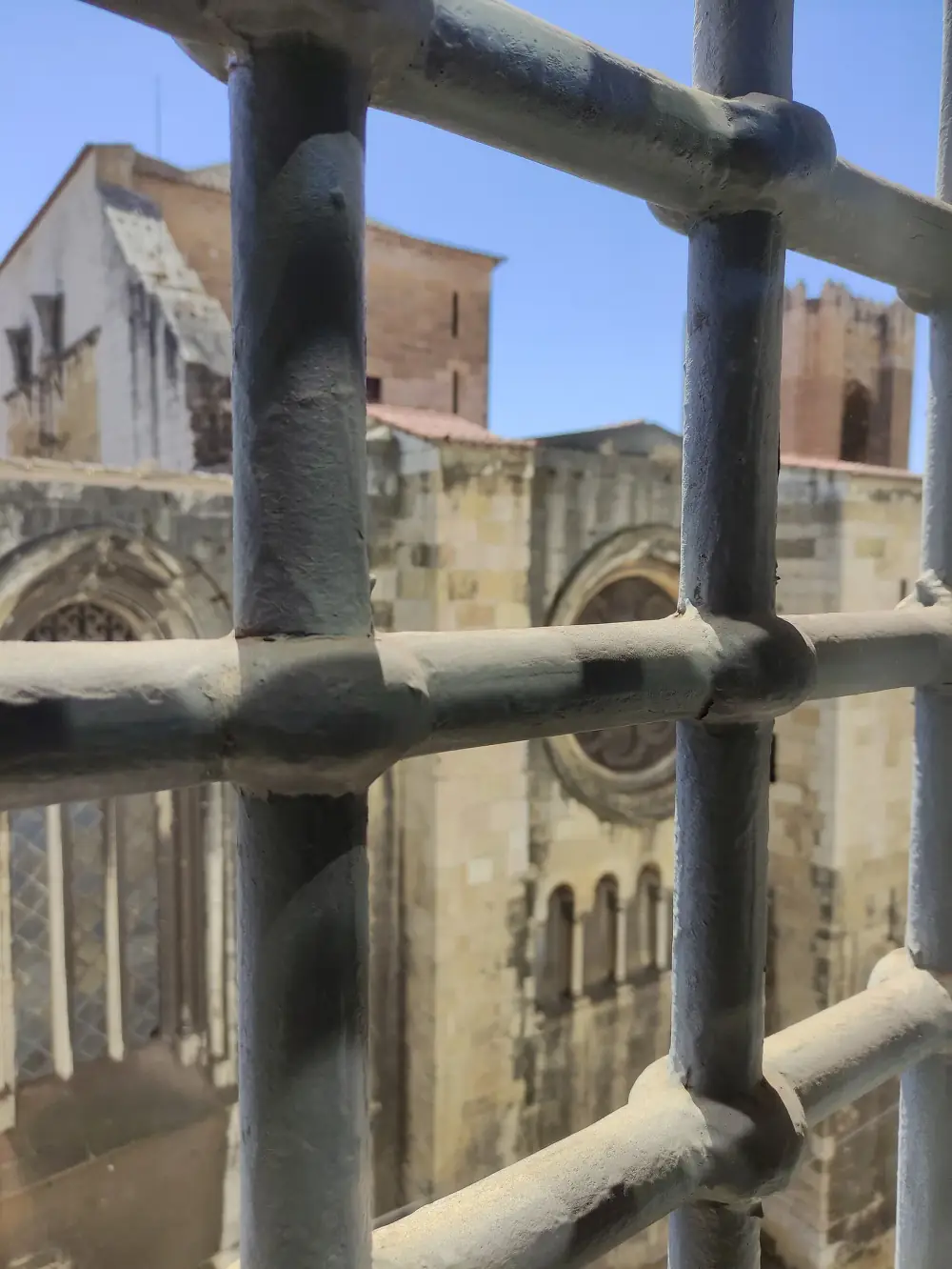 Thick grey iron bars fill the foreground. Through them, across a narrow gap, rises the pale stone wall of a medieval church with a rose window, with a crenellated tower behind it against a blue sky.