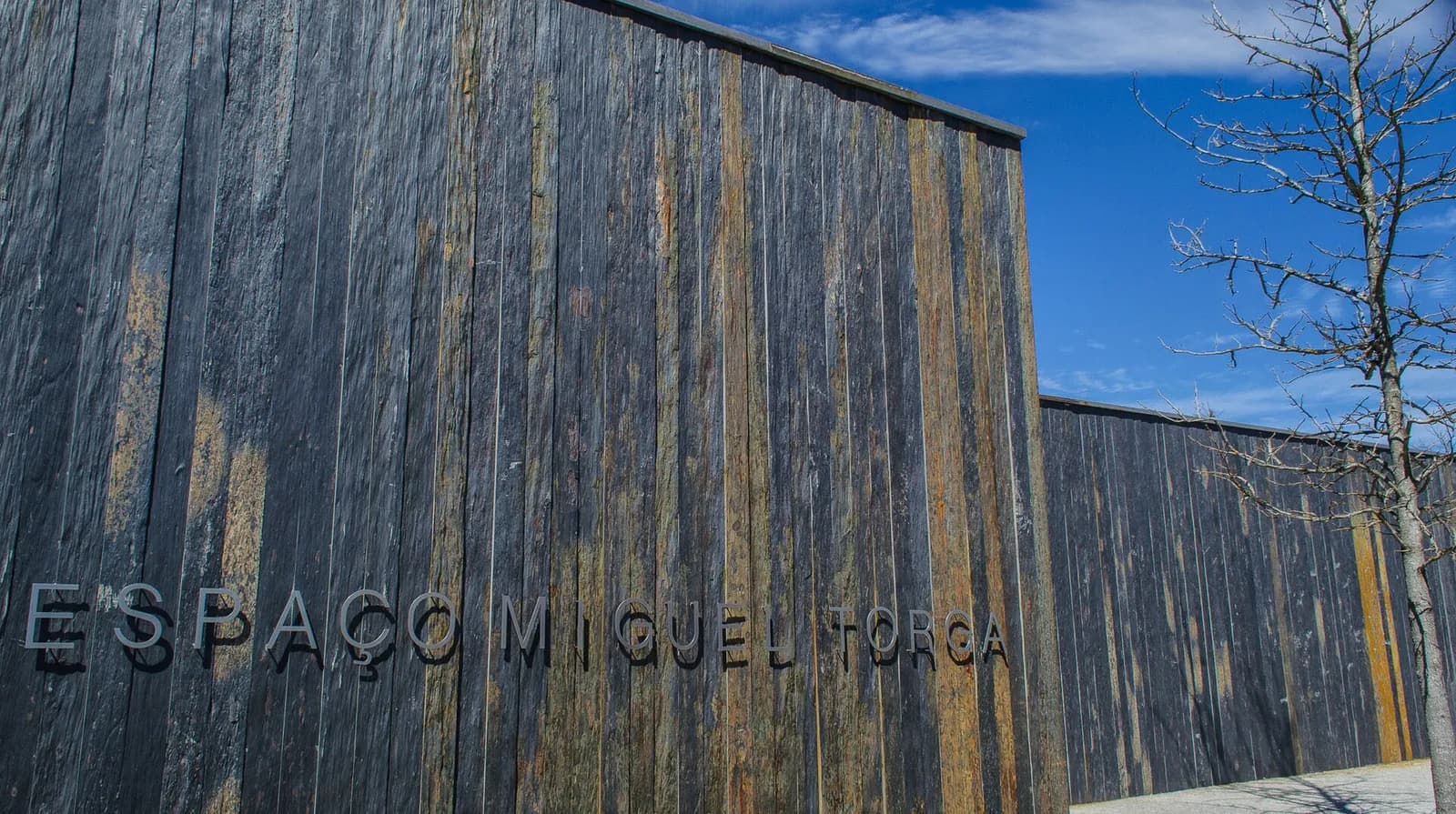A long wall of dark vertical slate slabs streaked with rust and grey, with the words Espaço Miguel Torga in metal letters across its lower part, under a blue sky with a bare tree to the right.