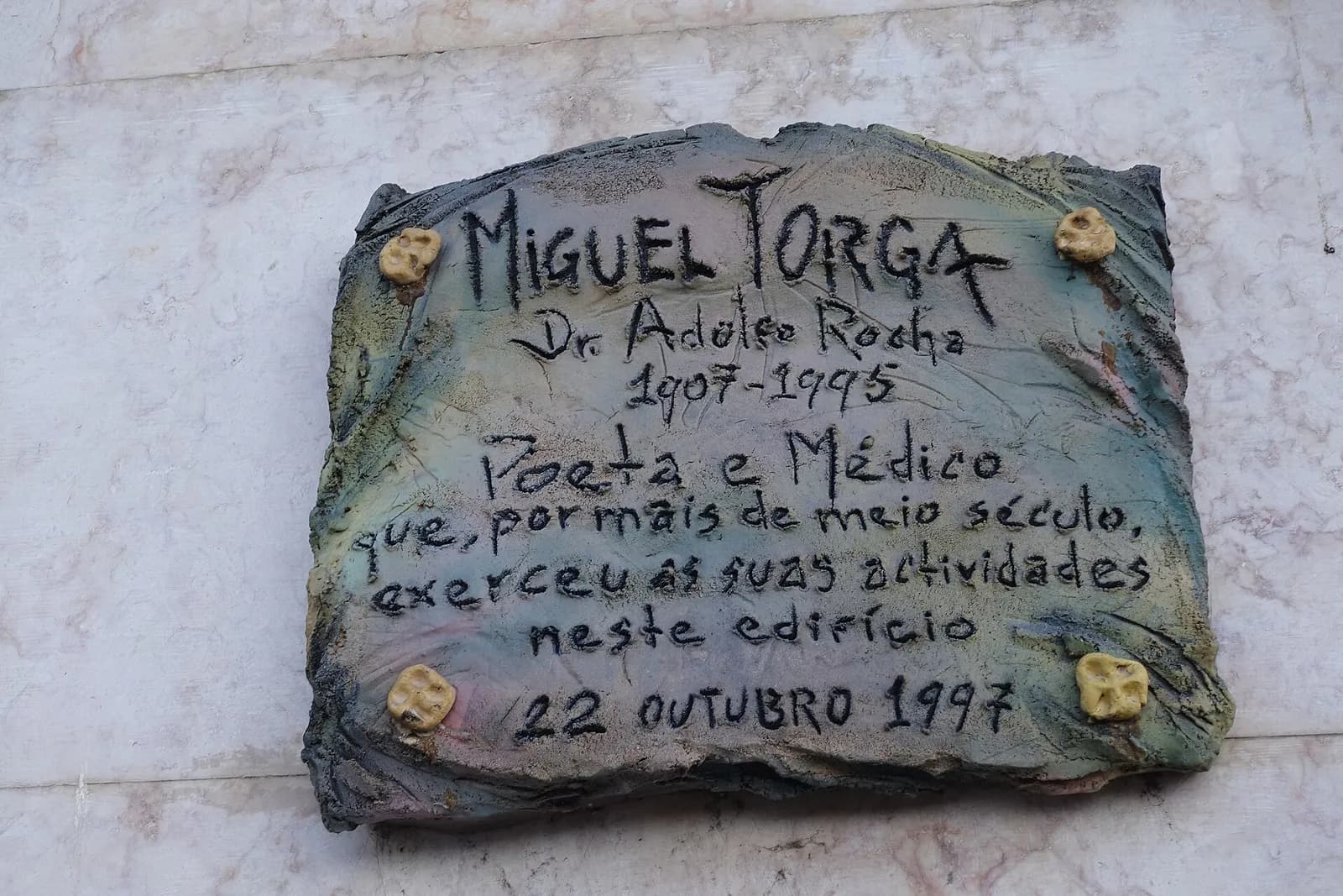 A rough-edged ceramic plaque in grey, green and pink glazes fixed to a pale marble wall, with hand-lettered text in black and four small yellow knobs at the corners.