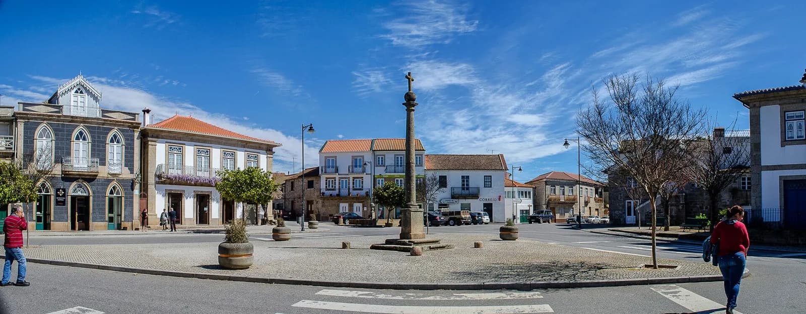A wide village square under a blue sky. A tall stone column topped by a cross stands on a cobbled island in the middle, with bare trees, white houses with orange-tiled roofs and a grey-fronted building with arched windows around it. A few people walk across the road.
