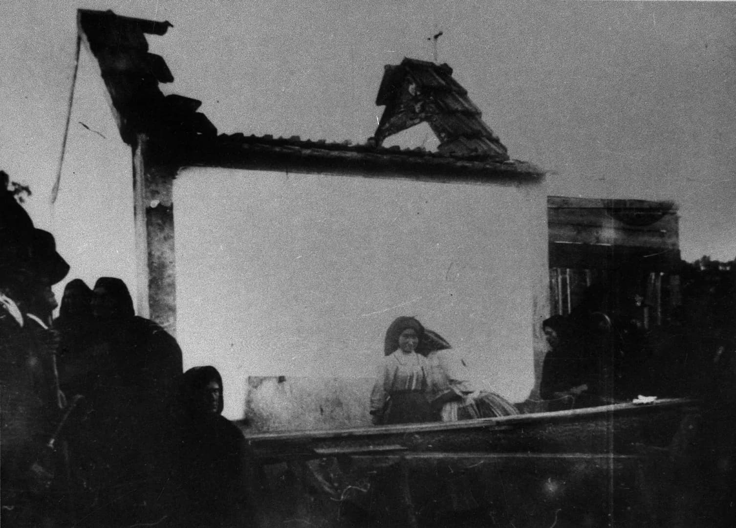A small whitewashed chapel with its roof torn open and its little bell gable knocked askew. A girl in a headscarf stands in front of the wall, and people in dark shawls stand to one side.