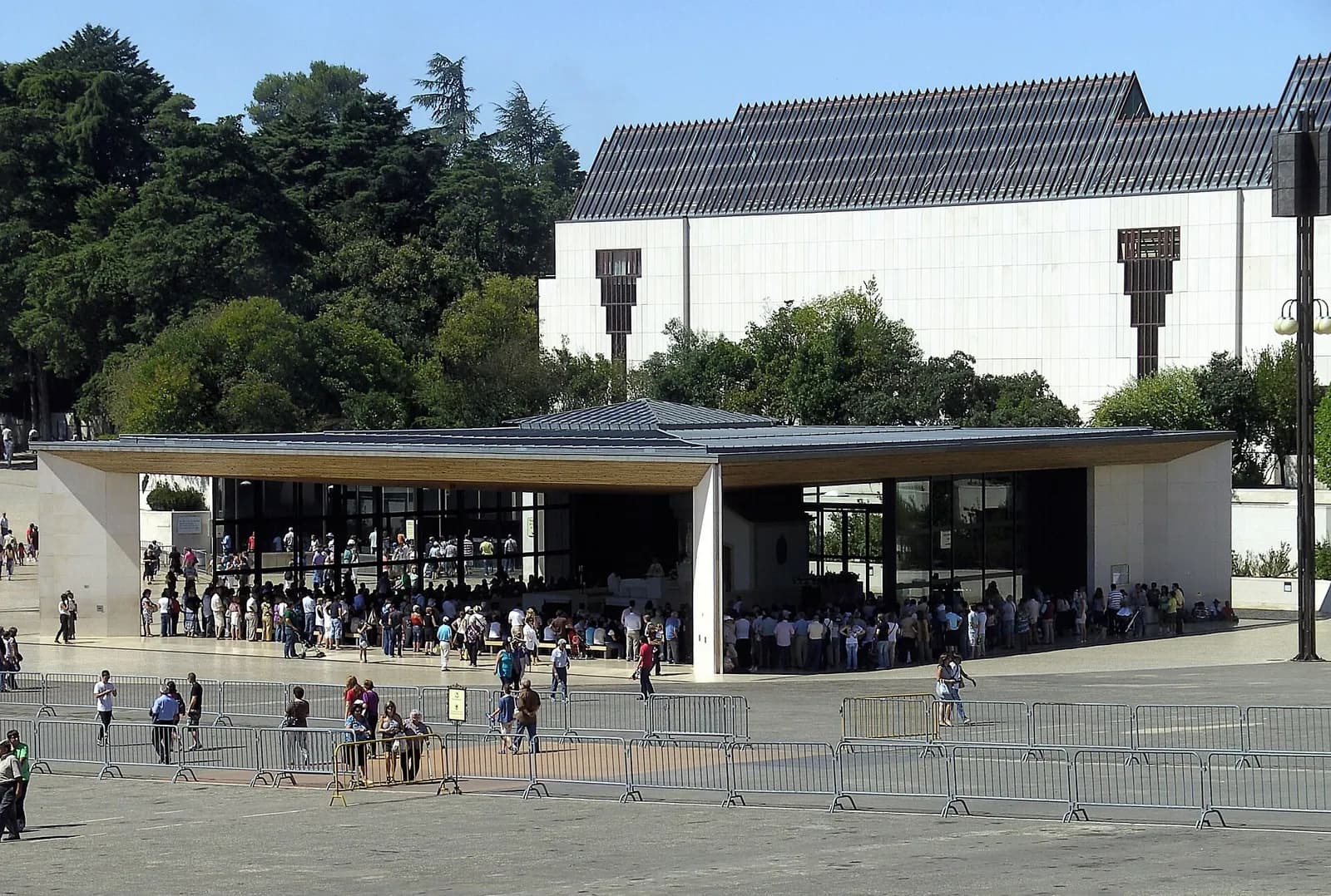 A low modern open-sided porch with a flat roof and glass walls, with pilgrims gathered inside and queuing outside, in front of a large white building and trees.