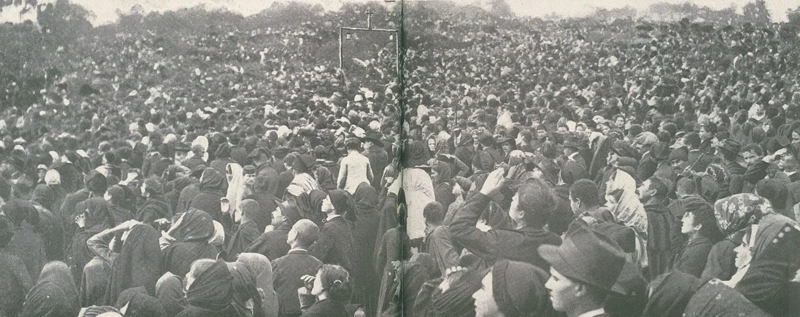 A dense crowd of men and women in dark clothes and headscarves fills a field to the horizon, many shading their eyes and looking up. In the middle distance a rough wooden frame hung with two lanterns and topped by a small cross rises above their heads.