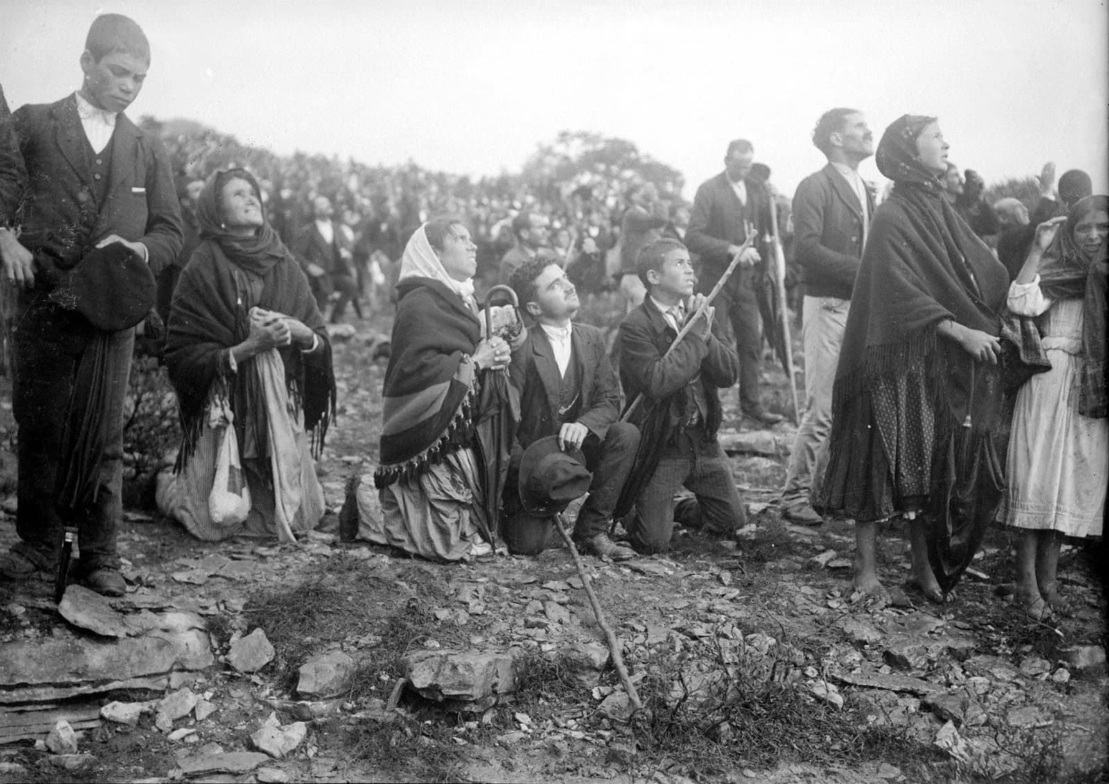 Men and women, several kneeling on stony ground with umbrellas, sticks and hats in hand, gaze upwards; a dense crowd stretches behind them.