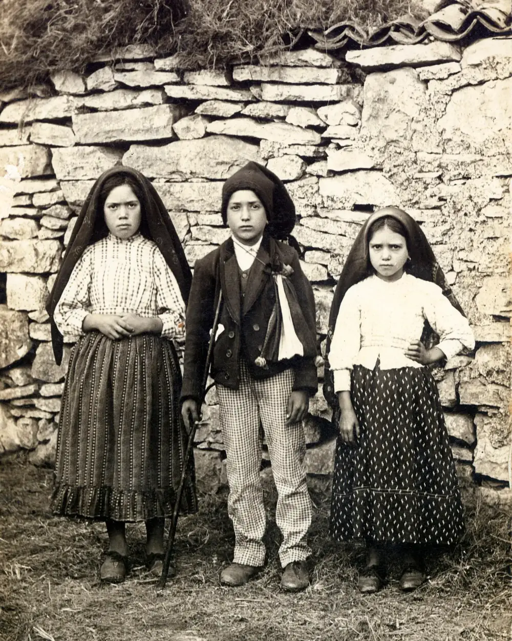 Three children stand in front of a dry-stone wall: a girl in a headscarf and striped skirt, a boy in a cap and jacket holding a stick, and a younger girl in a headscarf and dotted skirt.