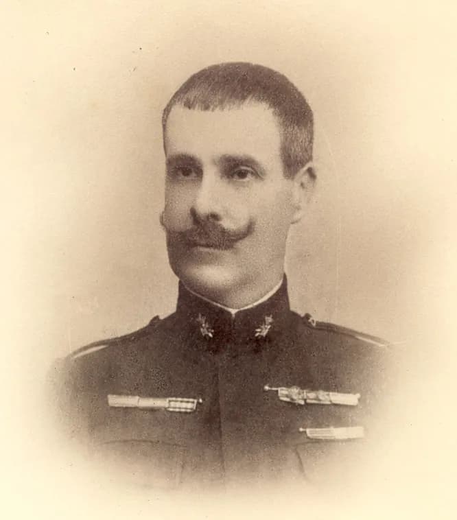 Studio portrait of a man with close-cropped hair and a large curled moustache, wearing a dark high-collared military tunic with medal ribbons.