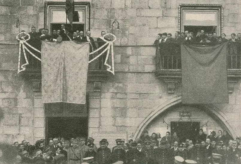Men in dark suits and hats crowd two balconies of a stone town hall hung with draped cloths and a flag, while officers in peaked caps and a crowd fill the square below.
