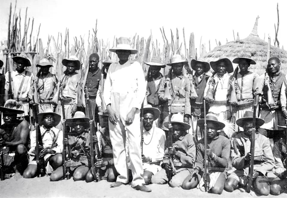 A tall man in a white suit and wide-brimmed hat stands among some two dozen men holding rifles, many in hats and bandoliers, some kneeling, in front of a palisade of wooden poles and a thatched hut.