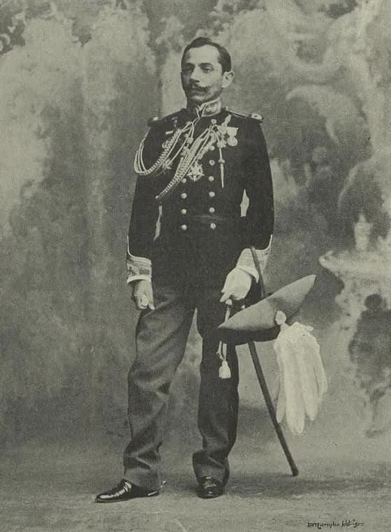 Full-length studio portrait of a moustached officer in a dark dress uniform with braid and medals, wearing white gloves and holding a plumed hat and a sword.