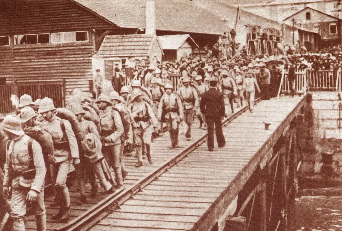 Columns of soldiers in pith helmets and light uniforms, with rolled blankets and packs on their backs, walk along a wooden pier, watched by a crowd behind a railing.