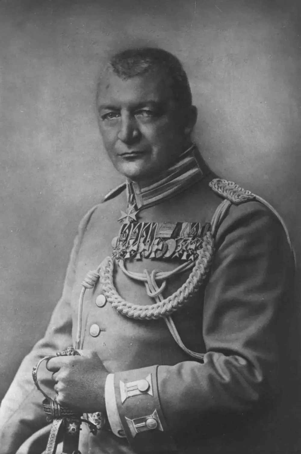 Head-and-shoulders portrait of a clean-shaven man with short grey hair in a light military tunic with braid, medal ribbons and a cross, his hands resting on a sword hilt.